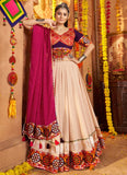 Woman in traditional outfit with colorful attire and pink dupatta in a decorated indoor setting.