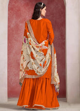 Woman wearing an orange traditional outfit with a floral dupatta against a red and white wall.