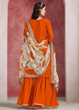 Woman wearing an orange traditional outfit with a floral dupatta against a red and white wall.