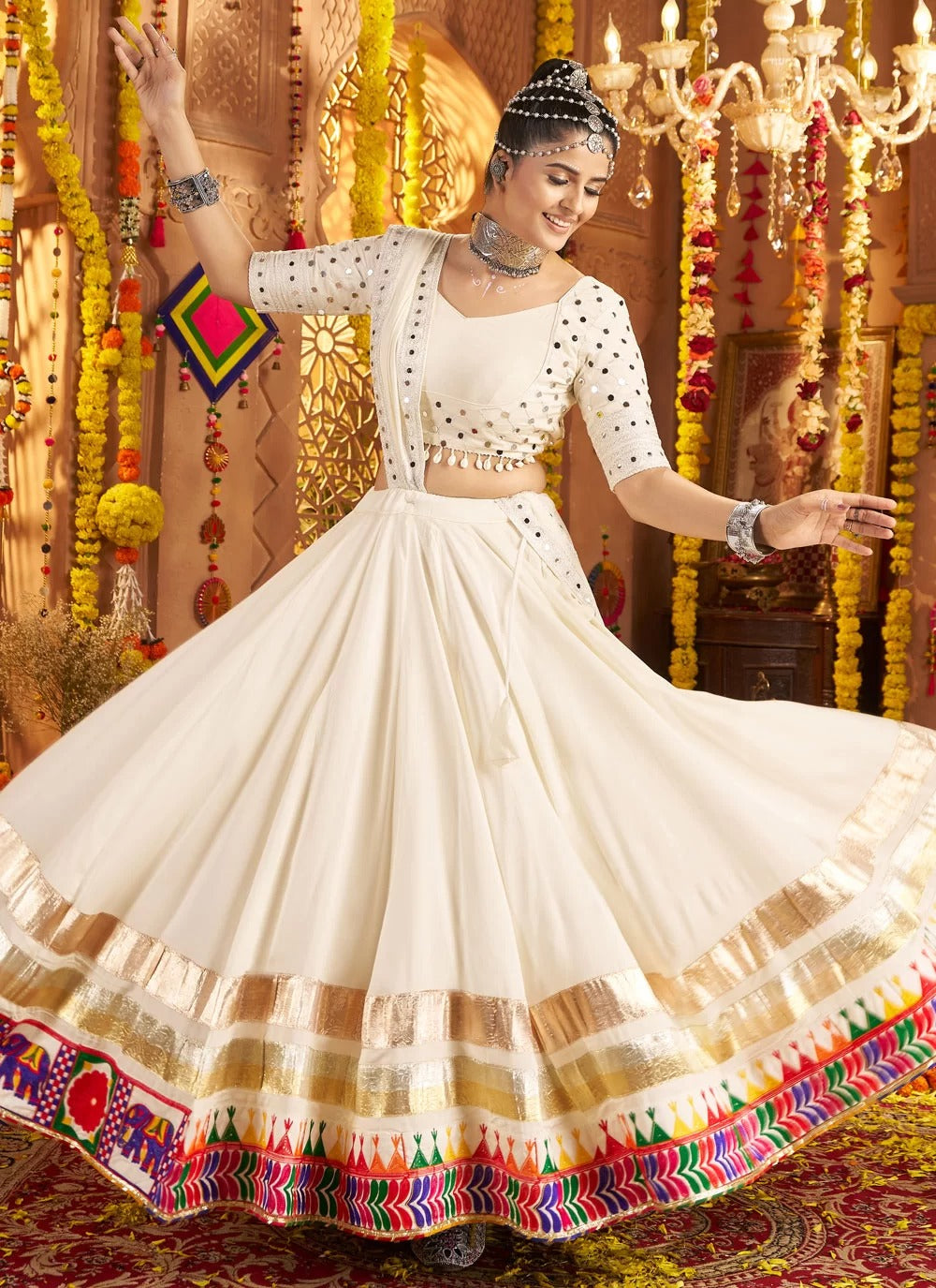 Woman in a traditional white and gold outfit with colorful border, standing in front of decorative background.
