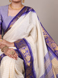Woman wearing a white saree with blue border and gold jewelry against a gray background