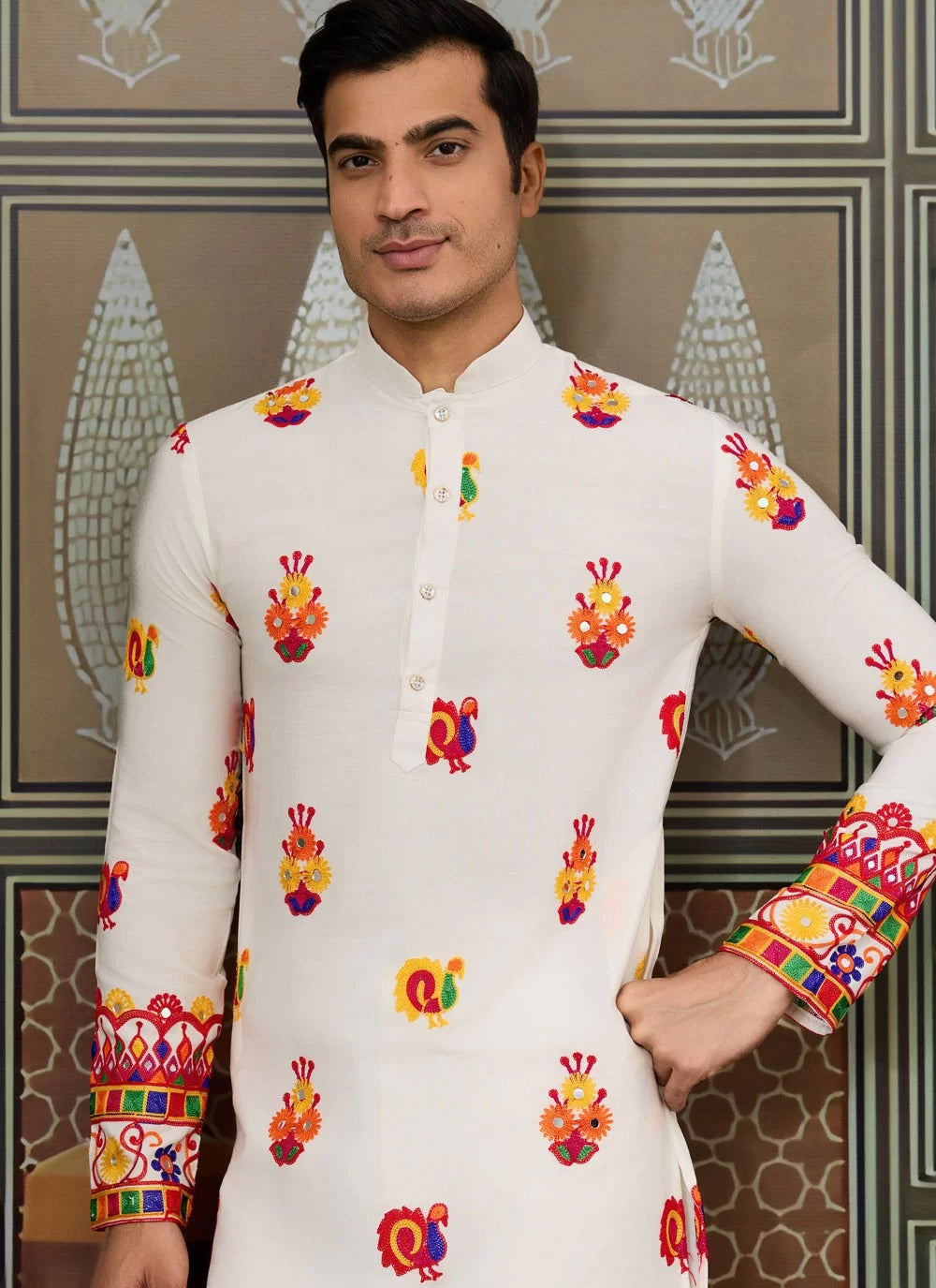 Man wearing a white kurta with colorful patterns against a decorative wall.