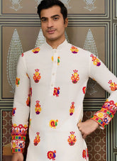 Man wearing a white kurta with colorful patterns against a decorative wall.