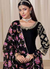 Woman wearing a black traditional outfit with floral patterns and jewelry against a beige curtain background