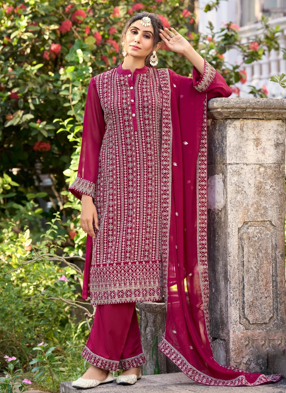 Woman in a traditional maroon and white embroidered outfit standing outdoors with greenery.