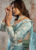 Woman wearing a light blue saree with intricate patterns against a textured wall.