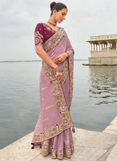 Woman in a pink saree with intricate patterns standing by a body of water.