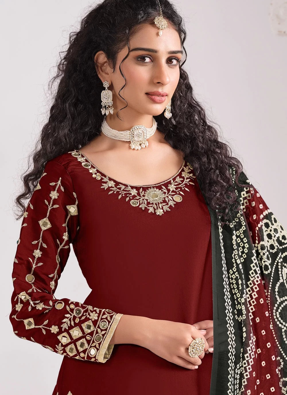 Woman wearing a maroon traditional outfit with gold embroidery and jewelry against a plain background