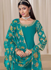 Woman wearing a teal embroidered traditional outfit with gold details against a neutral background