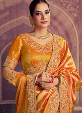 Woman wearing a traditional orange and yellow saree with intricate patterns and jewelry against a decorative background.