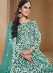 A woman wearing a mint green traditional embroidered outfit with white floral lace patterns, a matching dupatta, and ornate gold jewelry, posed against a warm beige background.