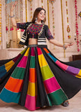 Woman in a colorful traditional outfit with a multicolored skirt and black top, standing in a decorated room.
