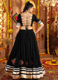 Woman wearing a black traditional outfit with intricate designs in a decorated indoor setting.