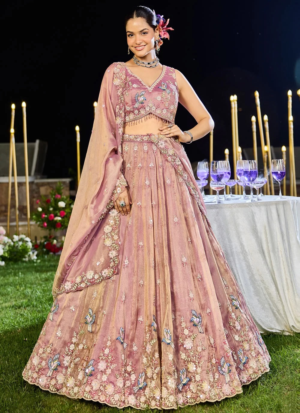 Woman in a pink embroidered traditional outfit at an outdoor event with candles and flowers.