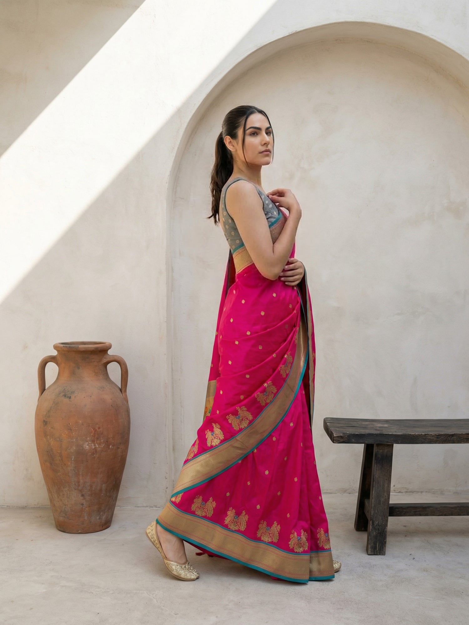 Woman in a pink saree standing in a rustic setting with a vase and bench.