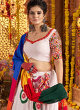 Woman in traditional outfit with colorful blouse and saree, holding a green bag, against a decorated background.