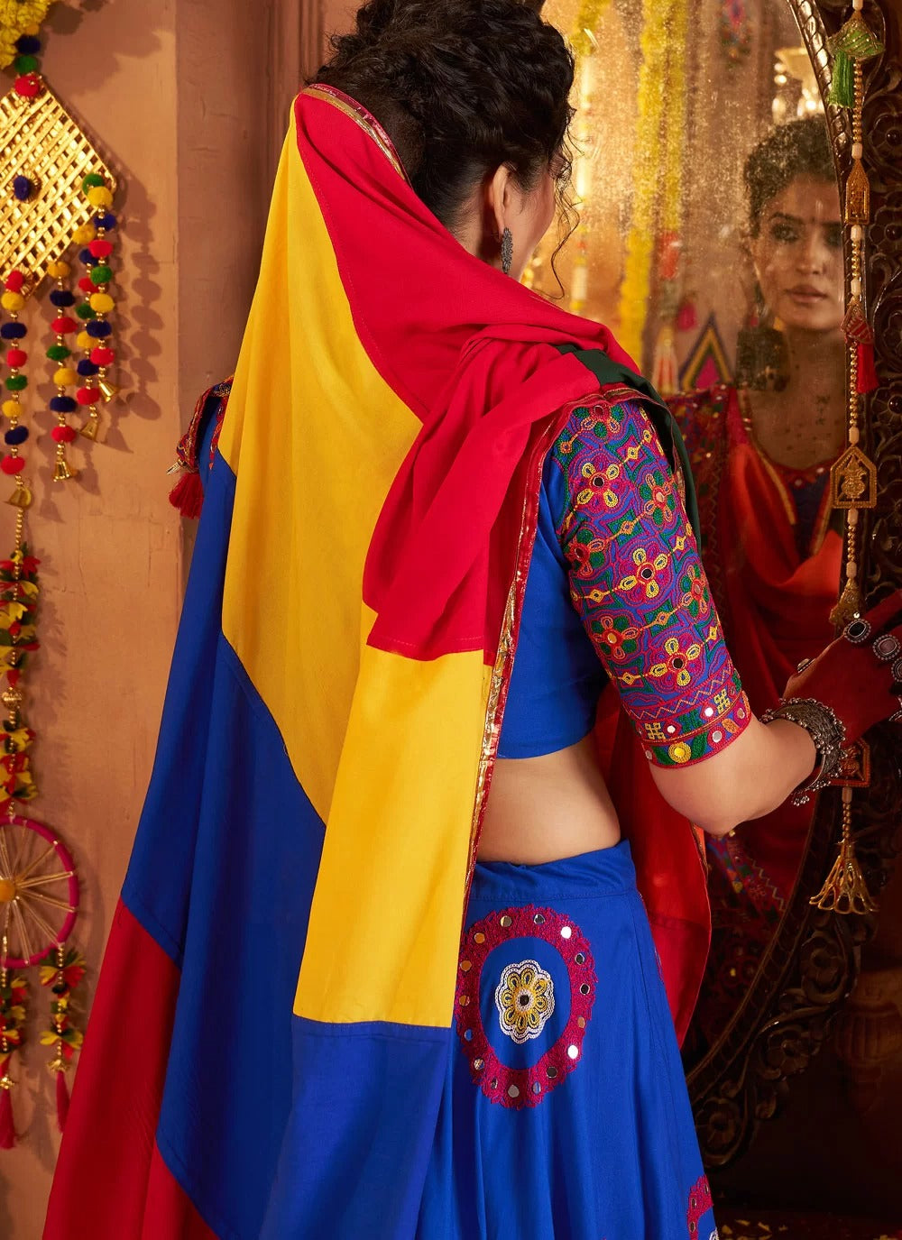 Woman in colorful traditional outfit with a red, yellow, and blue scarf, standing in front of a mirror.