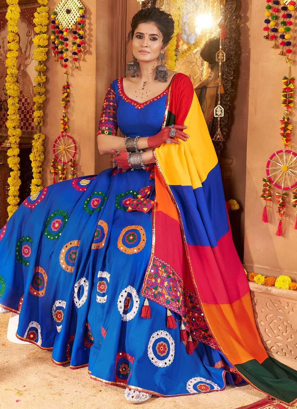 Woman in a colorful traditional outfit with a vibrant blue saree and multicolored dupatta against a decorated background.