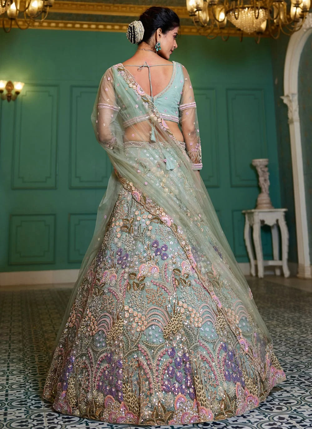 Woman in a traditional embroidered lehenga in an elegant room with green walls and chandeliers.