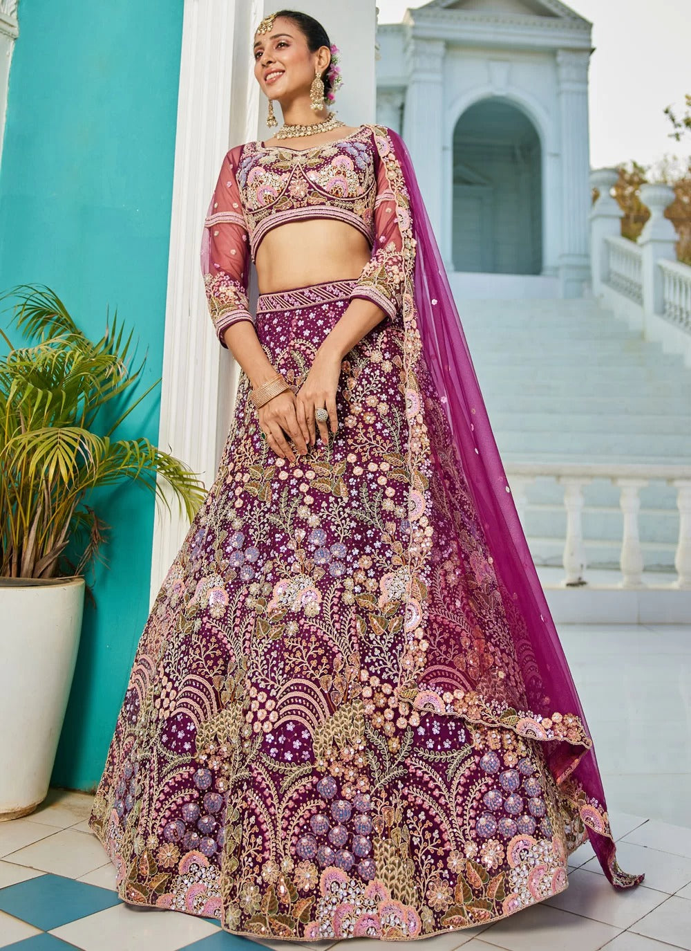 Woman in a traditional embroidered outfit with a pink dupatta standing in front of a blue wall.