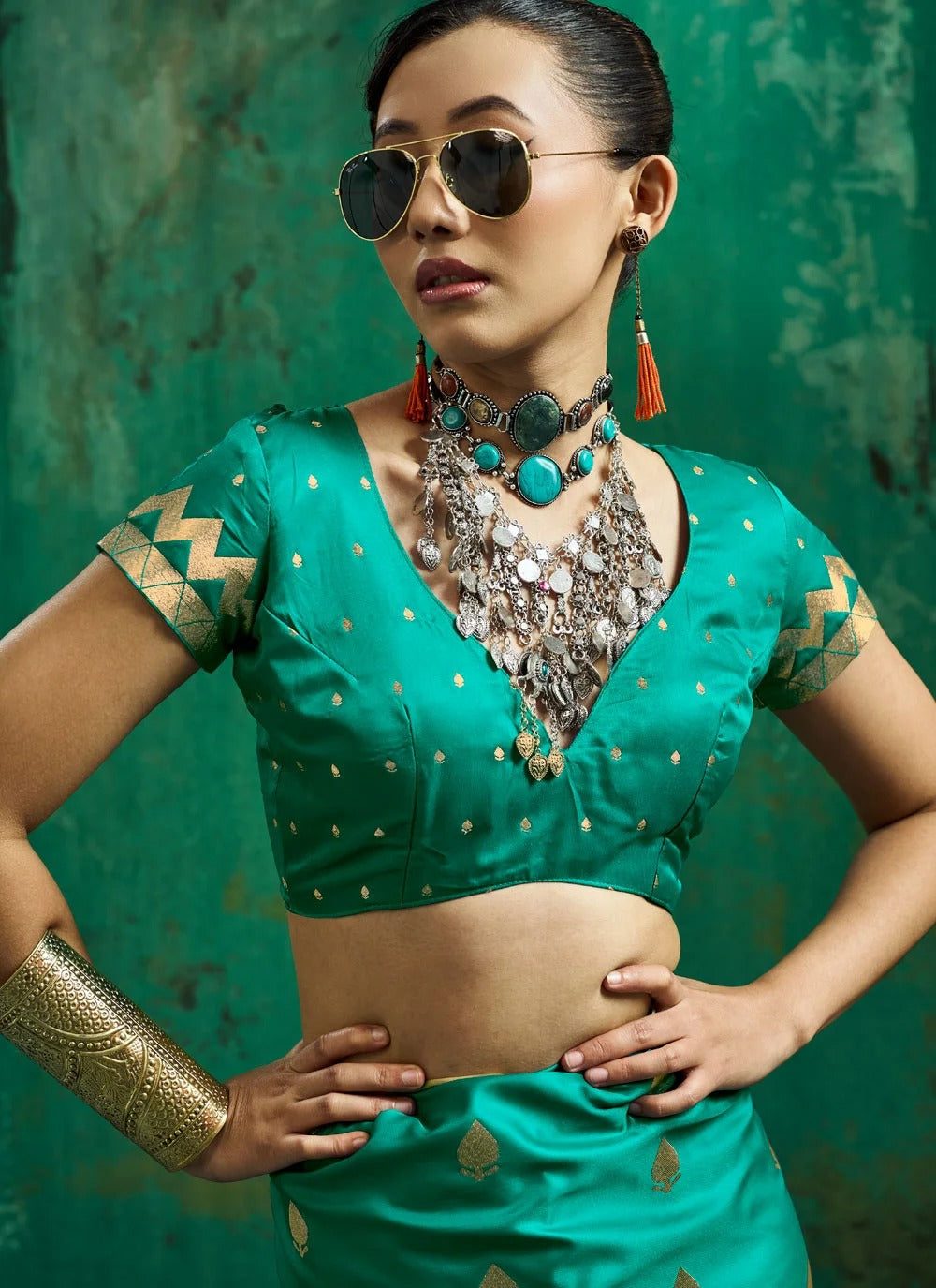A woman wearing a teal green traditional Indian blouse and matching skirt with gold motifs, accessorized with a large silver necklace, gold bangle, and sunglasses, posed against a textured green backdrop.