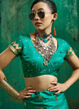 A woman wearing a teal green traditional Indian blouse and matching skirt with gold motifs, accessorized with a large silver necklace, gold bangle, and sunglasses, posed against a textured green backdrop.