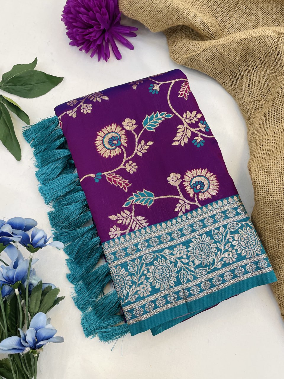 Purple fabric with floral patterns and blue tassels on a white surface with flowers and burlap.