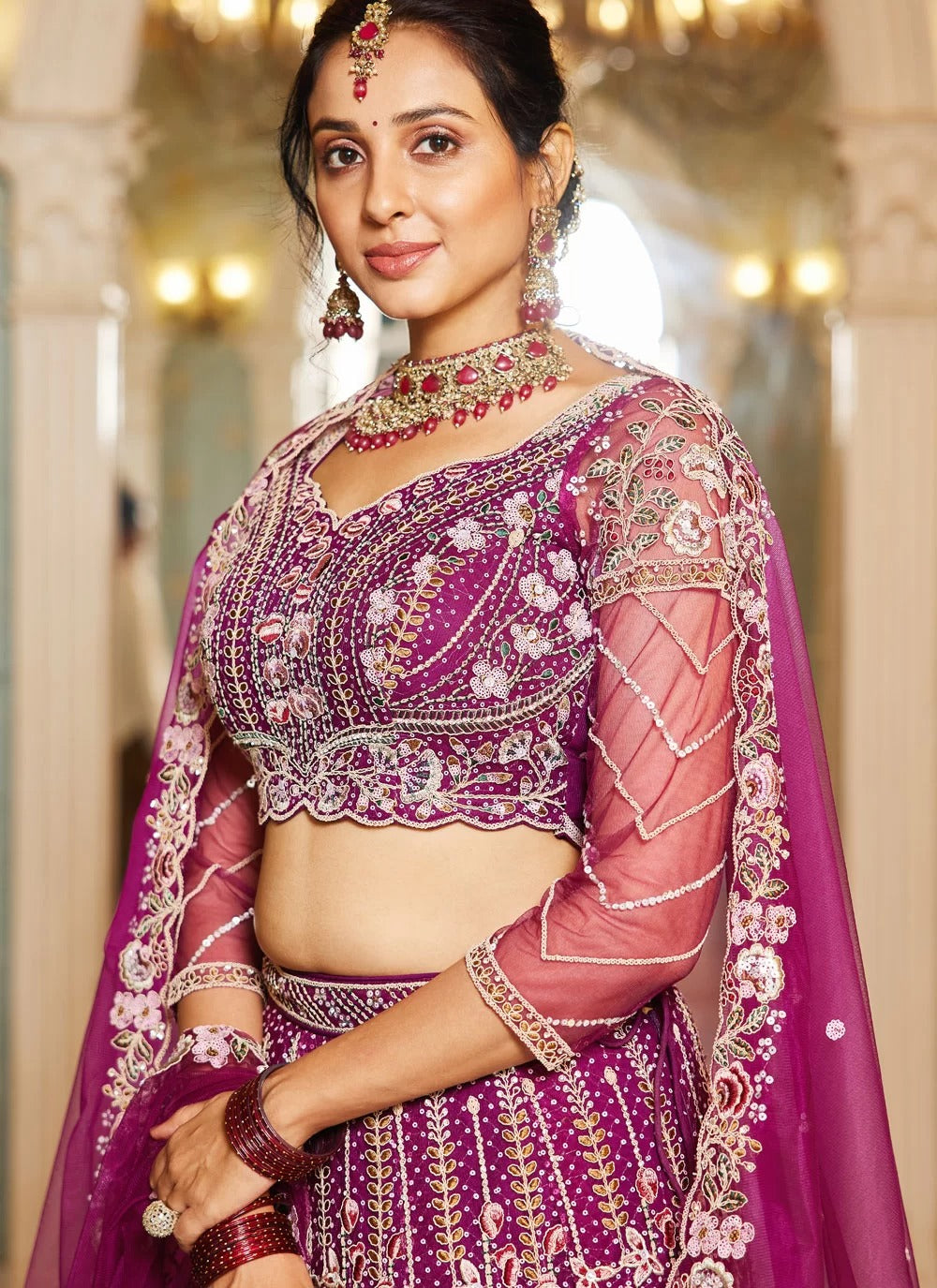 Woman in a traditional purple and pink embroidered outfit with jewelry in an ornate setting.