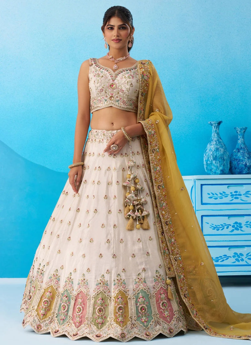 A woman models a traditional Indian lehenga choli set in a studio setting with a blue background. The outfit consists of a cream-colored embroidered skirt (lehenga), a matching embroidered crop top (choli), and a sheer mustard yellow dupatta draped over her shoulder.