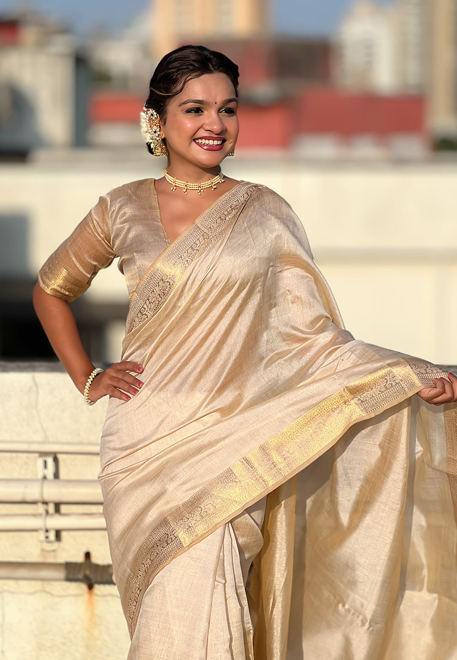 Woman in a beige saree with gold border standing outdoors.