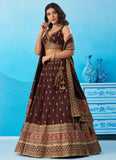 A woman models a traditional Indian lehenga choli suit in dark brown with gold and red metallic embroidery, set against a blue studio backdrop with decorative blue vases and a cabinet.