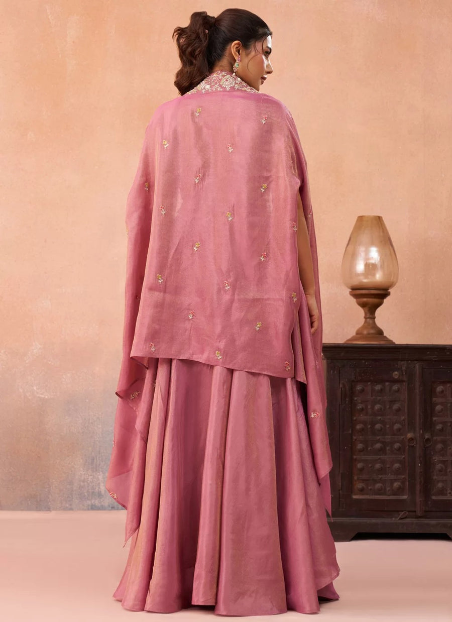 Woman in a pink traditional outfit standing against a peach-colored wall with a lamp and wooden cabinet.