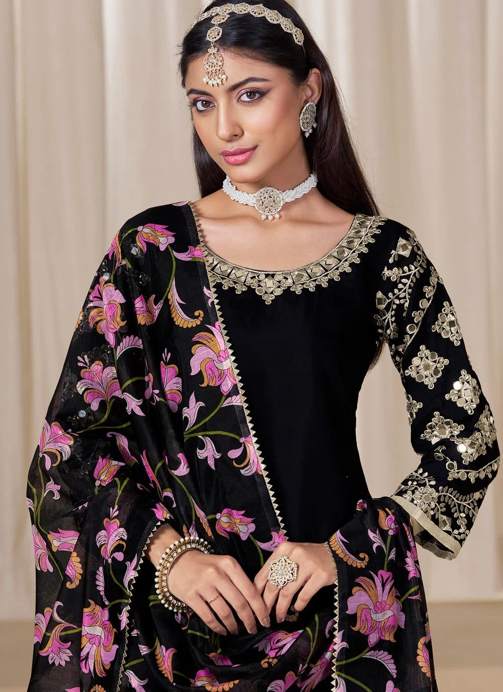 Woman wearing a black traditional outfit with floral patterns and jewelry against a beige curtain background