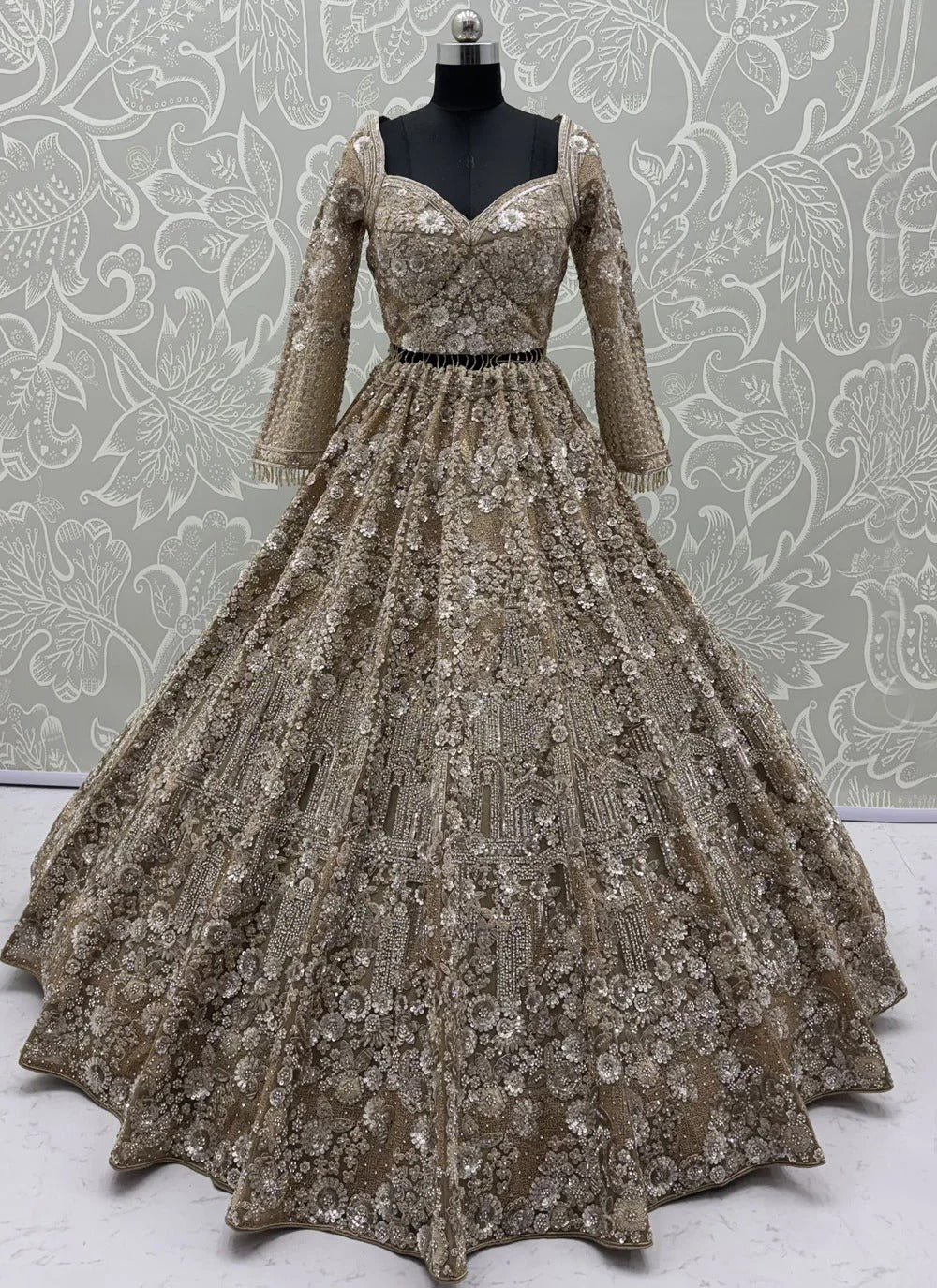 A floor-length ball gown displayed on a black mannequin, featuring a sweetheart neckline, three-quarter sleeves, and an intricate floral lace and sequin pattern in champagne and gold tones, set against a patterned wallpaper background.