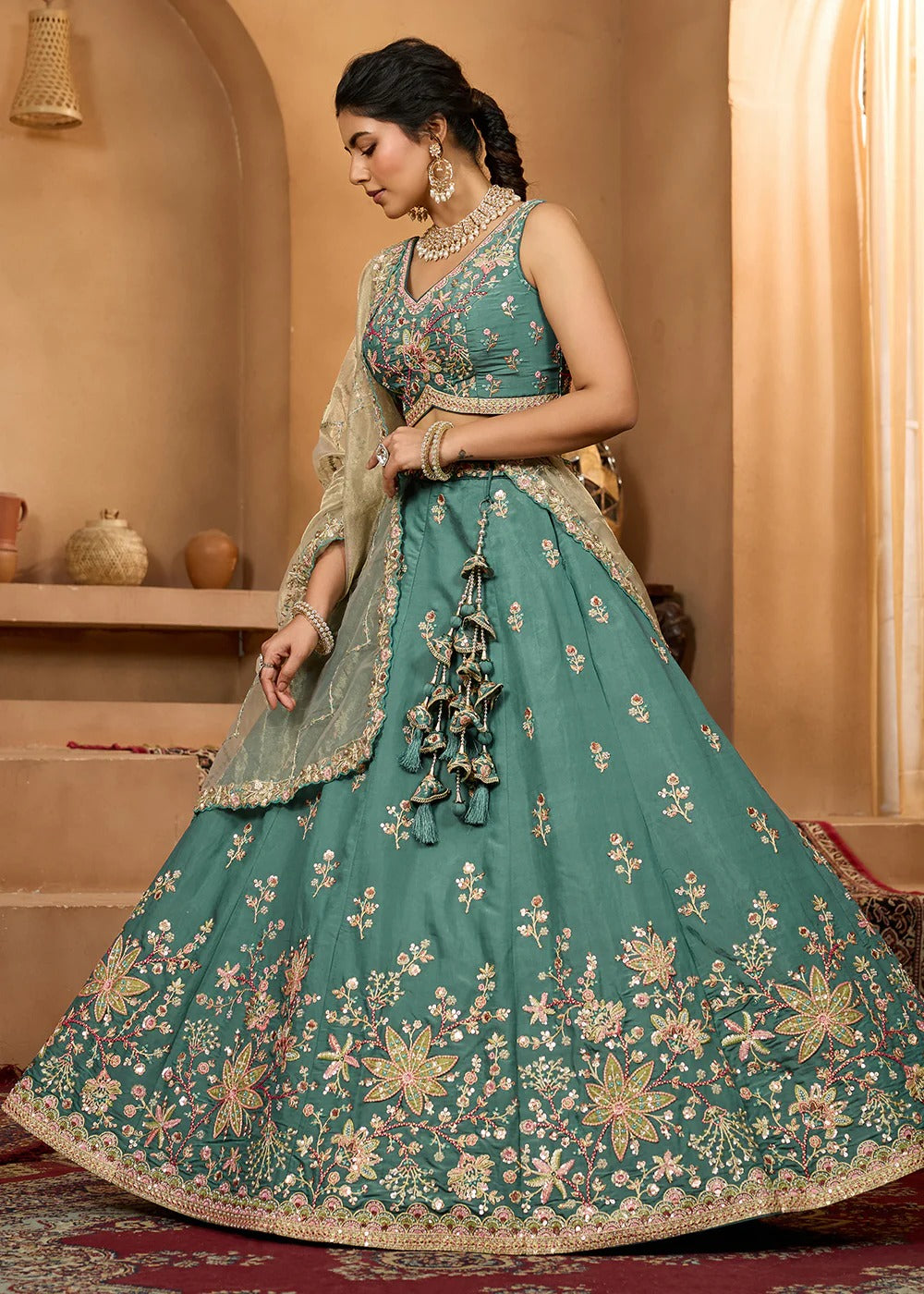 Woman in a teal embroidered traditional outfit with gold details in a warm-toned room.
