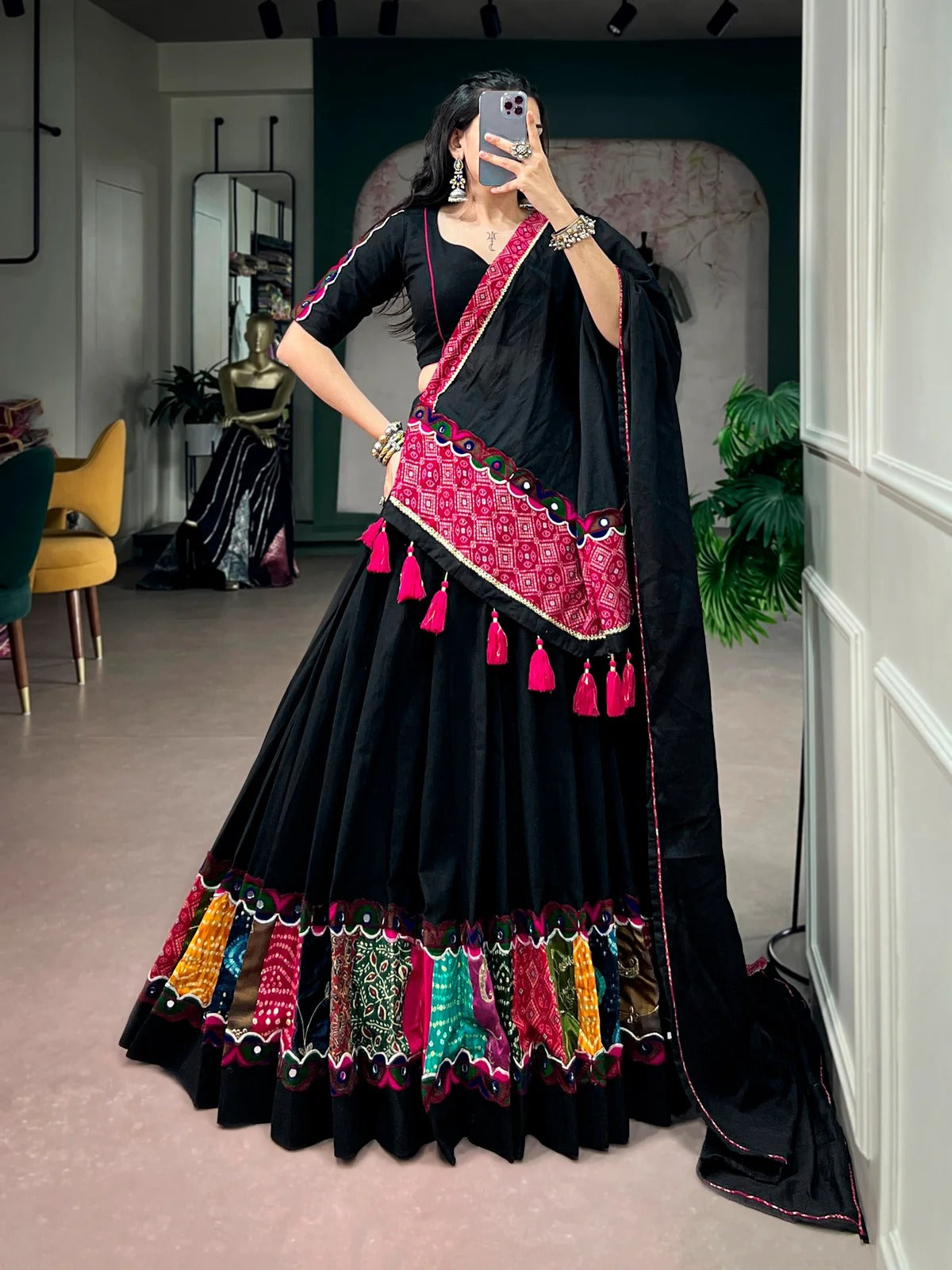 A woman takes a mirror selfie wearing a black traditional Indian lehenga choli with vibrant pink, red, and multicolor embroidered borders and tassels, set in an indoor boutique environment.