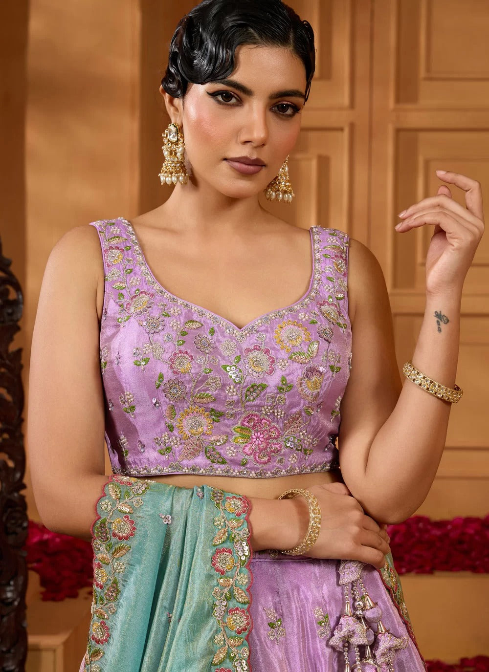 Woman wearing a traditional outfit with a purple blouse and green dupatta in a decorated room.