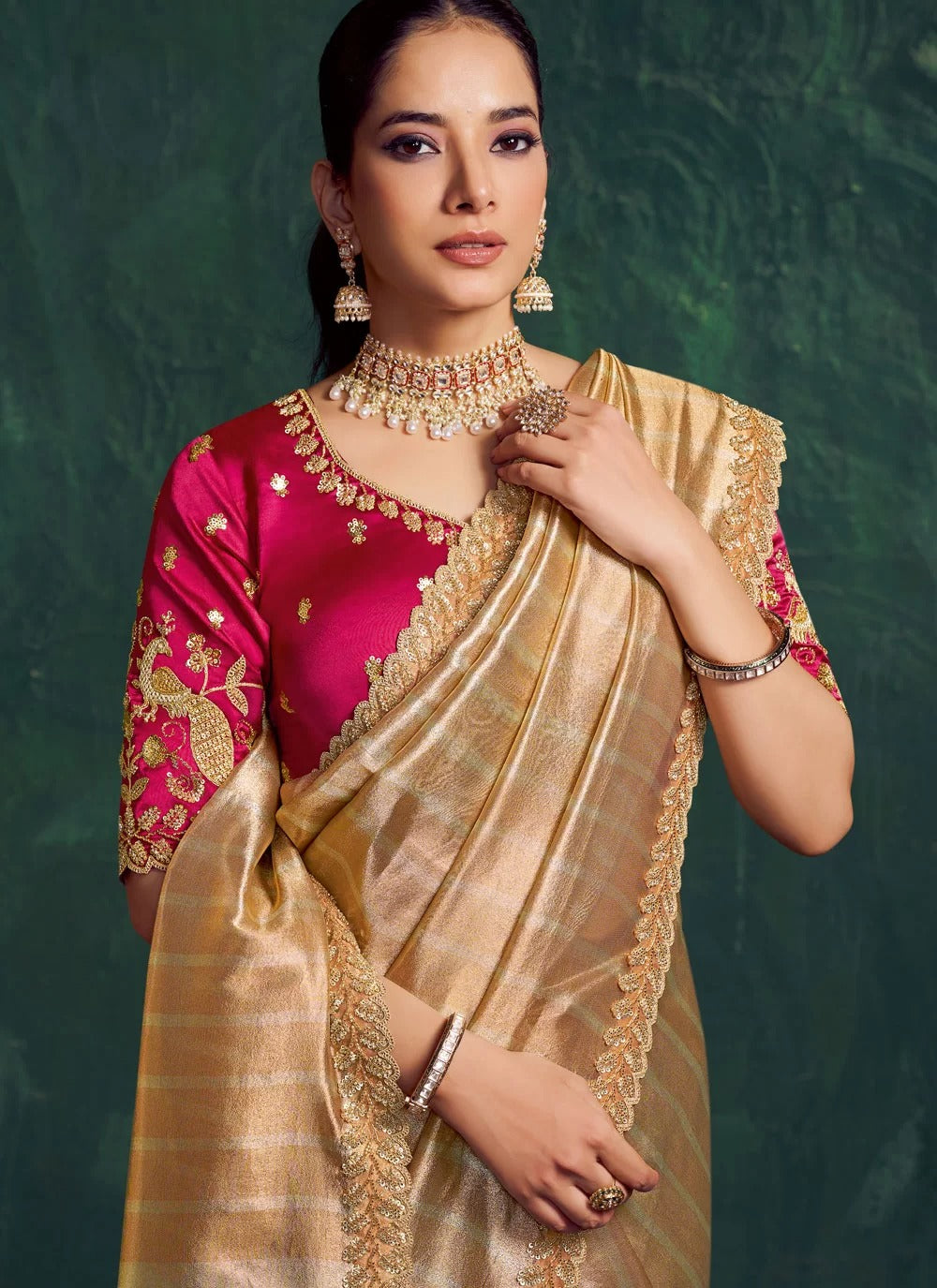 Woman wearing a traditional gold and pink saree with jewelry against a green background