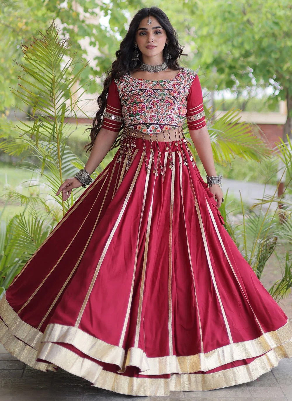 A woman wearing a traditional Indian red lehenga choli outfit with gold and multicolor embroidery, standing outdoors in front of green plants.