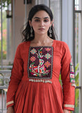 A woman wearing a terracotta orange-red Anarkali-style dress with colorful hand-embroidered floral and animal motifs on the chest and sleeve, set against a softly blurred indoor background.