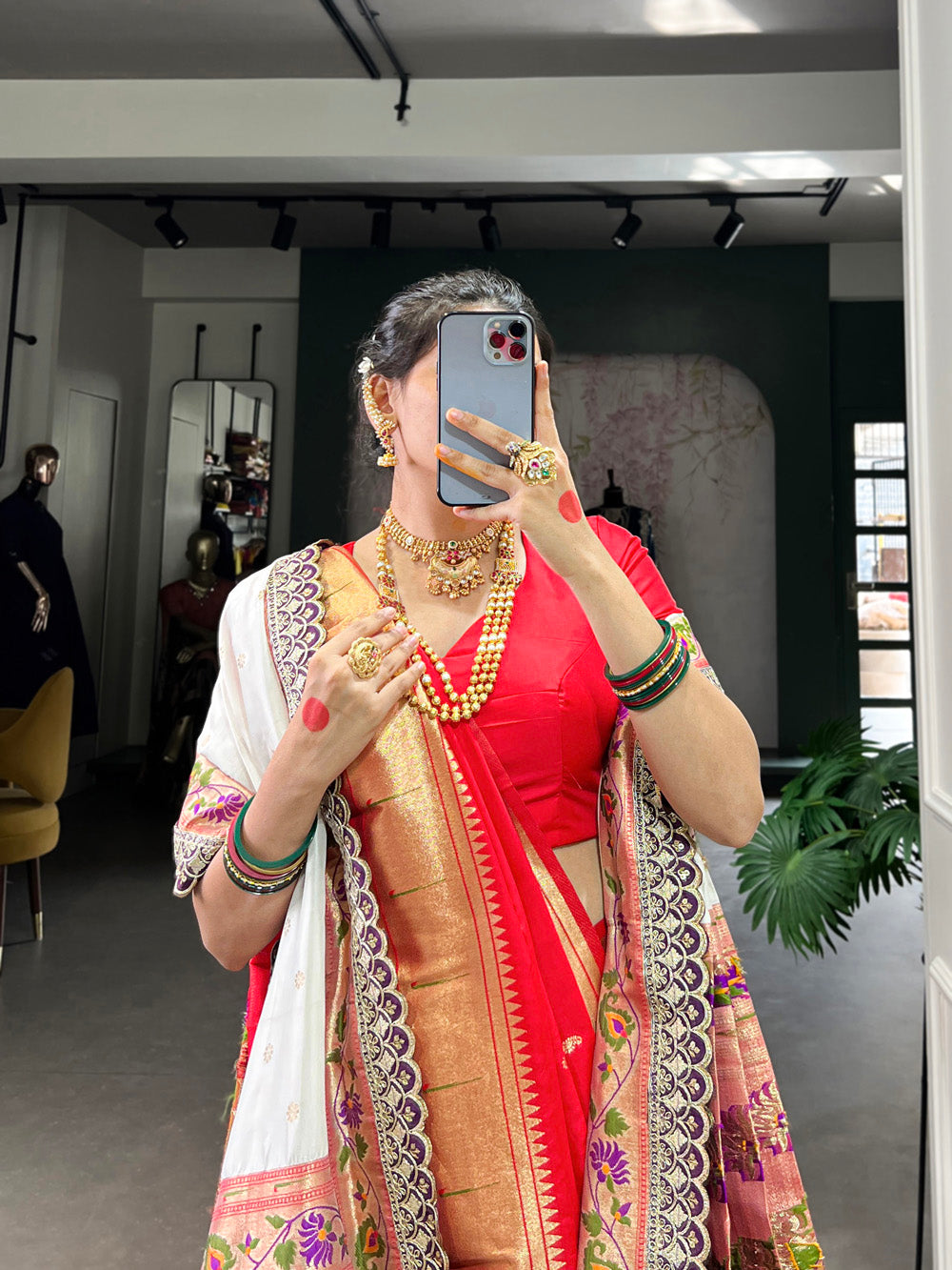 Woman in a red saree with gold jewelry taking a mirror selfie.