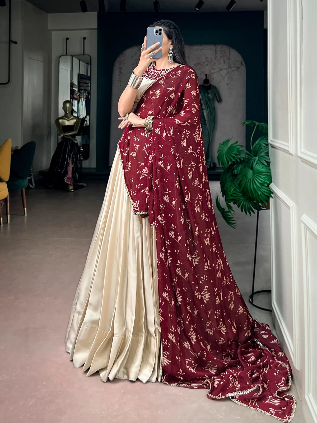 A woman takes a mirror selfie in a boutique wearing a traditional Indian salwar kameez outfit. The outfit features a deep maroon red dupatta and border with gold floral patterns, paired with a cream-colored satin body and skirt.