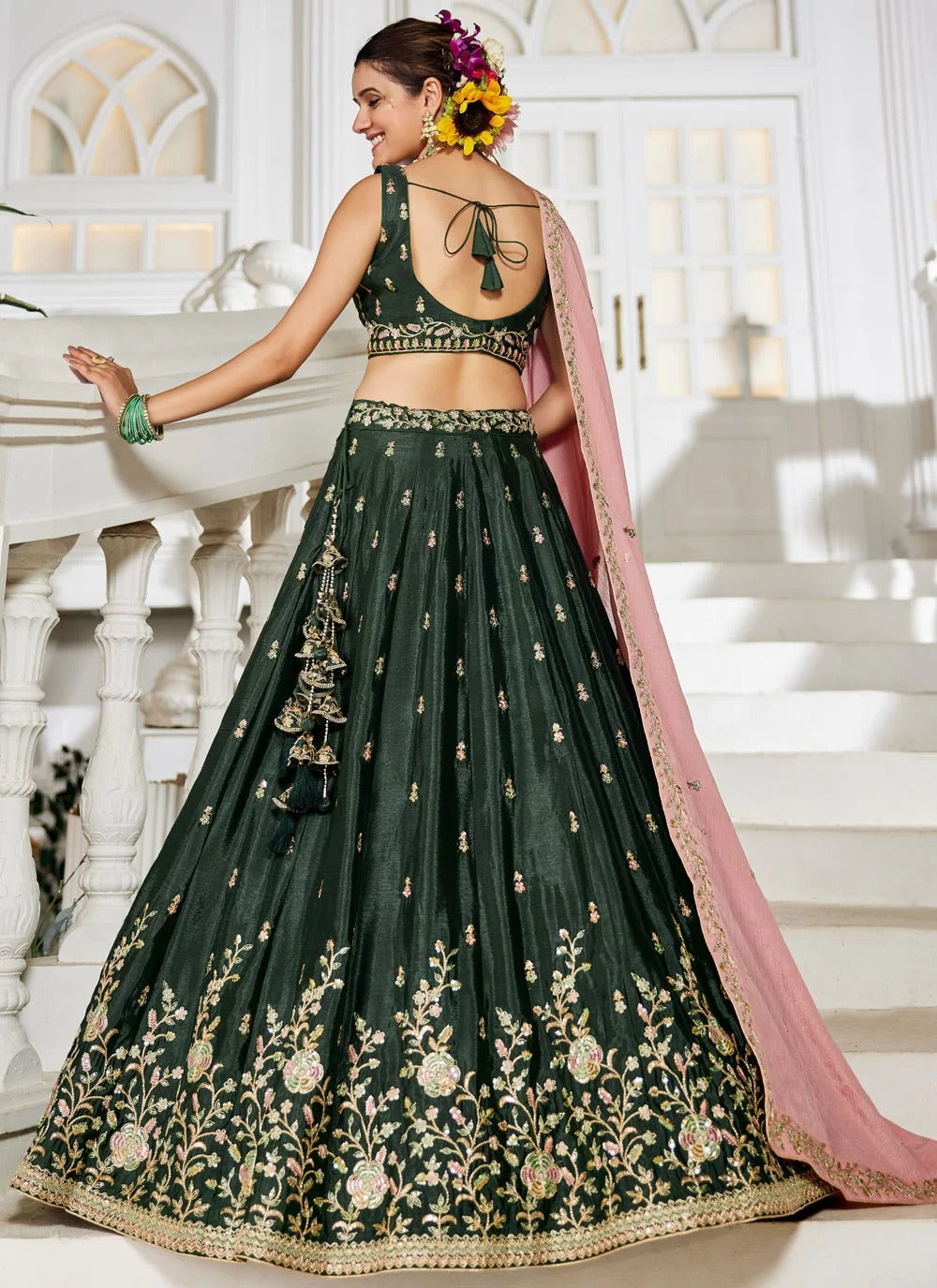 Woman in a dark green embroidered lehenga with a pink dupatta standing on a staircase.