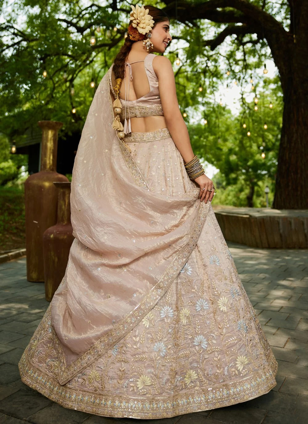 Woman in a traditional pink saree with floral decorations, standing outdoors with greenery in the background.