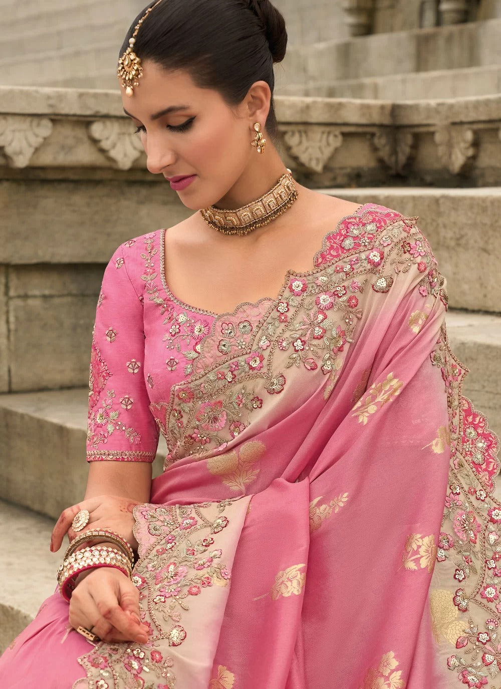 Woman wearing a pink saree with intricate embroidery sitting on steps.