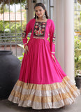 A woman models a bright pink traditional long dress with a heavily embroidered neckline and sleeve panels, featuring a tiered ruffled hem in beige, gold, and white metallic fabrics, set against a porch background.