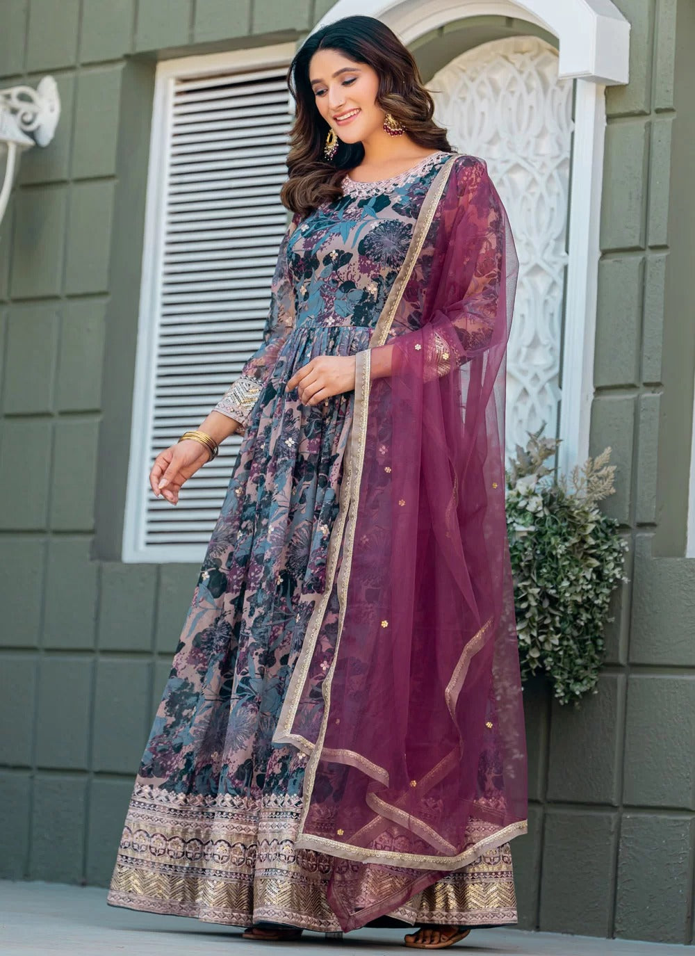 Woman wearing a traditional outfit with a floral patterned dress and purple dupatta in front of a gray building.