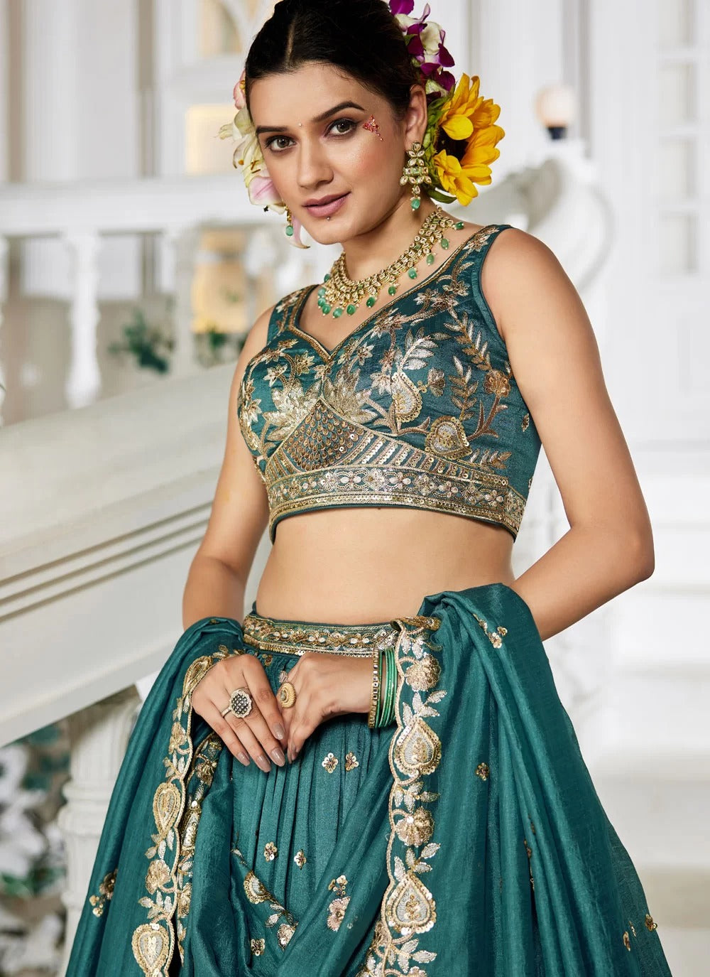 Woman in traditional green and gold embroidered outfit with floral hair accessories in a decorated indoor setting.