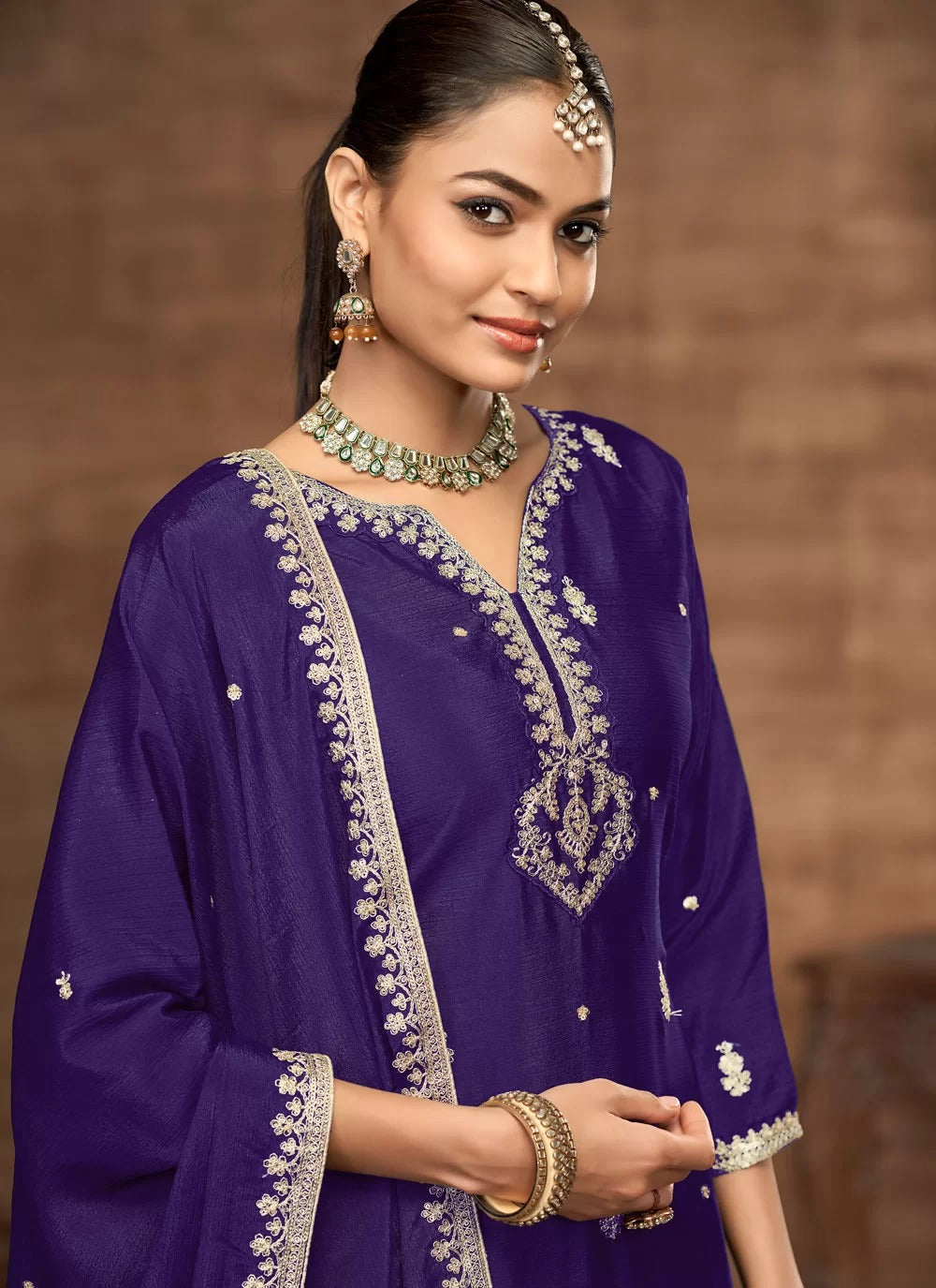 Woman wearing a traditional purple outfit with intricate designs and jewelry against a brown background