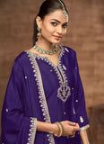 Woman wearing a traditional purple outfit with intricate designs and jewelry against a brown background